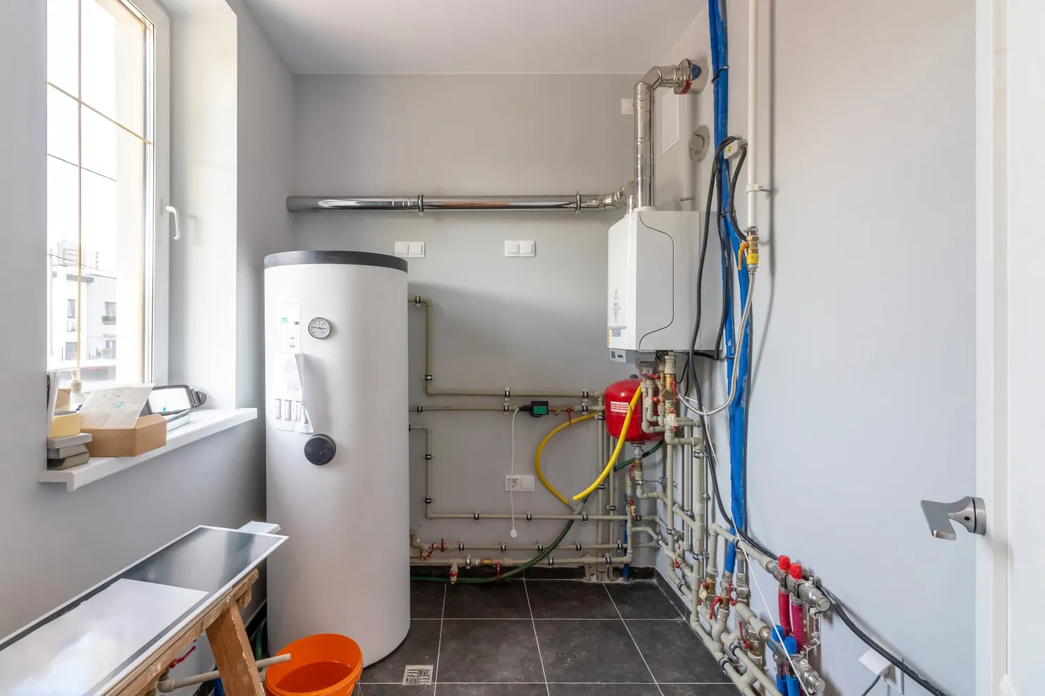 A hot water cylinder standing beside a wall-mounted boiler and its pipework in a plant room