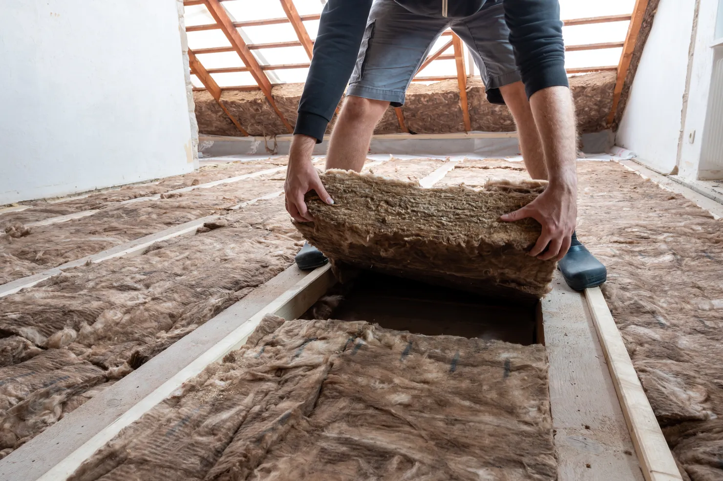 Installer laying a rock wool insulation batt between the floor joists of a loft, with insulation already fitted across the surrounding bays